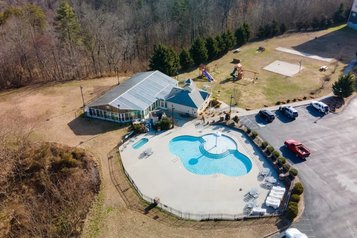 An inviting outdoor scene showing a large swimming pool surrounded by a recreational area, with playground equipment visible nearby. Children play, families gather, and the area is bathed in cheerful daylight, suggesting a joyful, energetic environment.