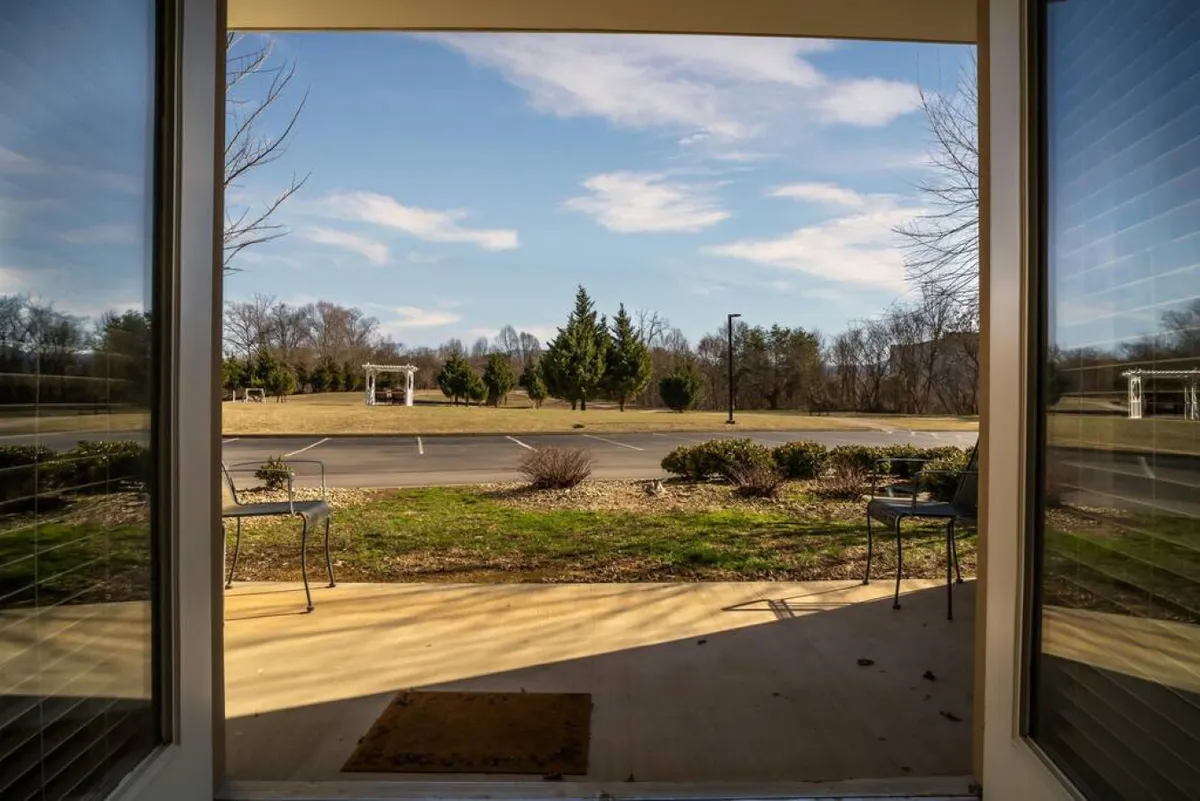 An inviting front porch with two chairs overlooks an open view of the landscape, featuring lush greenery and a blue sky. The scene evokes relaxation and social moments, hinting at the opportunity for guests to unwind outdoors after a lively night out.