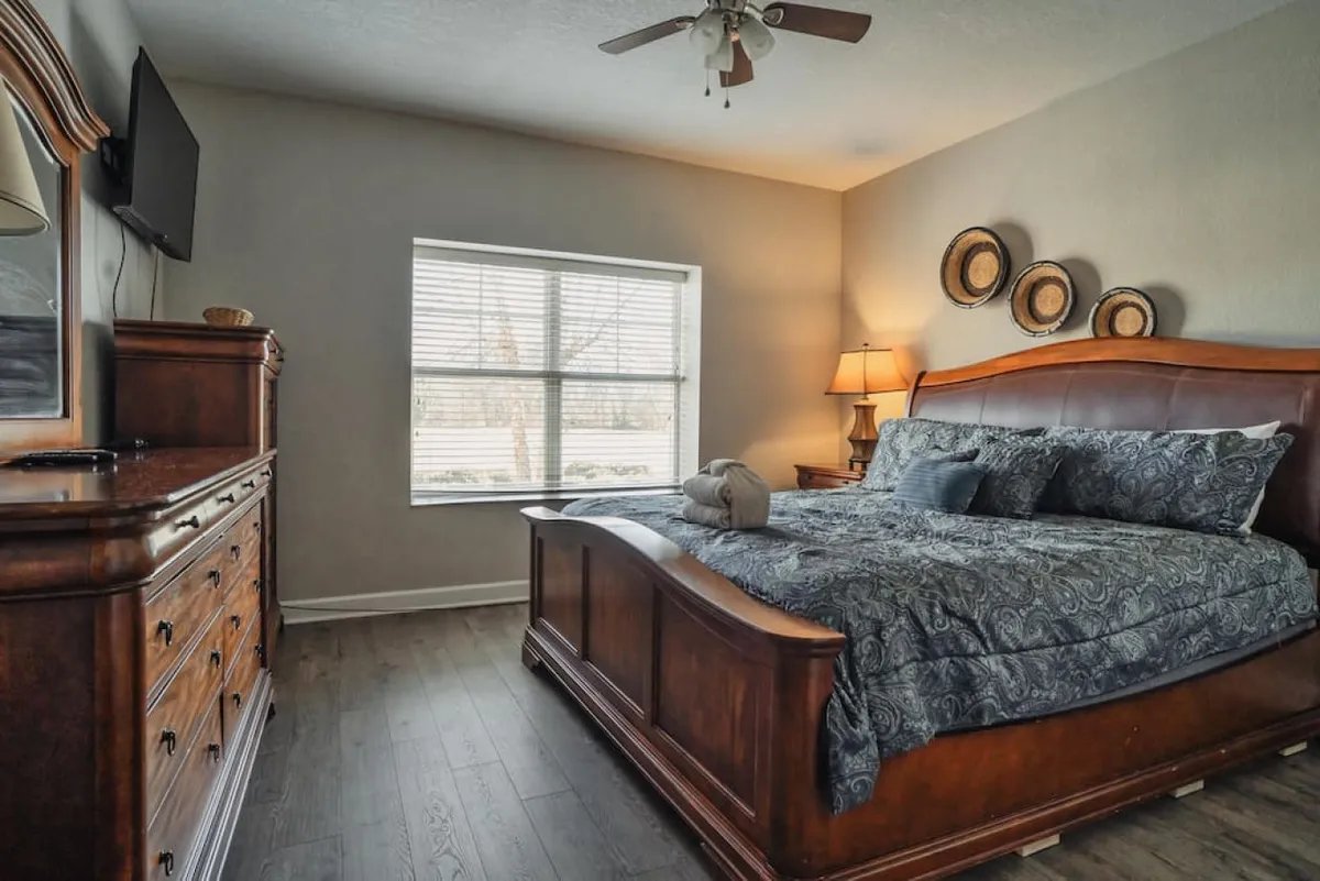 A spacious bedroom with a large wooden bed adorned with decorative pillows, a nightstand with a lamp, a dresser with a mirror, and a window letting in natural light. The overall atmosphere is calm and restful, hinting at comfort and privacy.