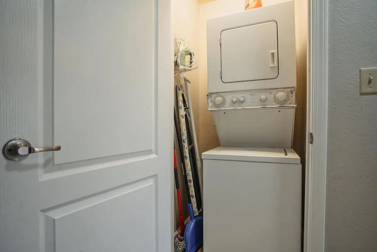 A compact laundry area featuring a stacked washer and dryer, with cleaning supplies and organized storage surrounding the appliances. The space is bright, efficient, and clearly focused on guest convenience.