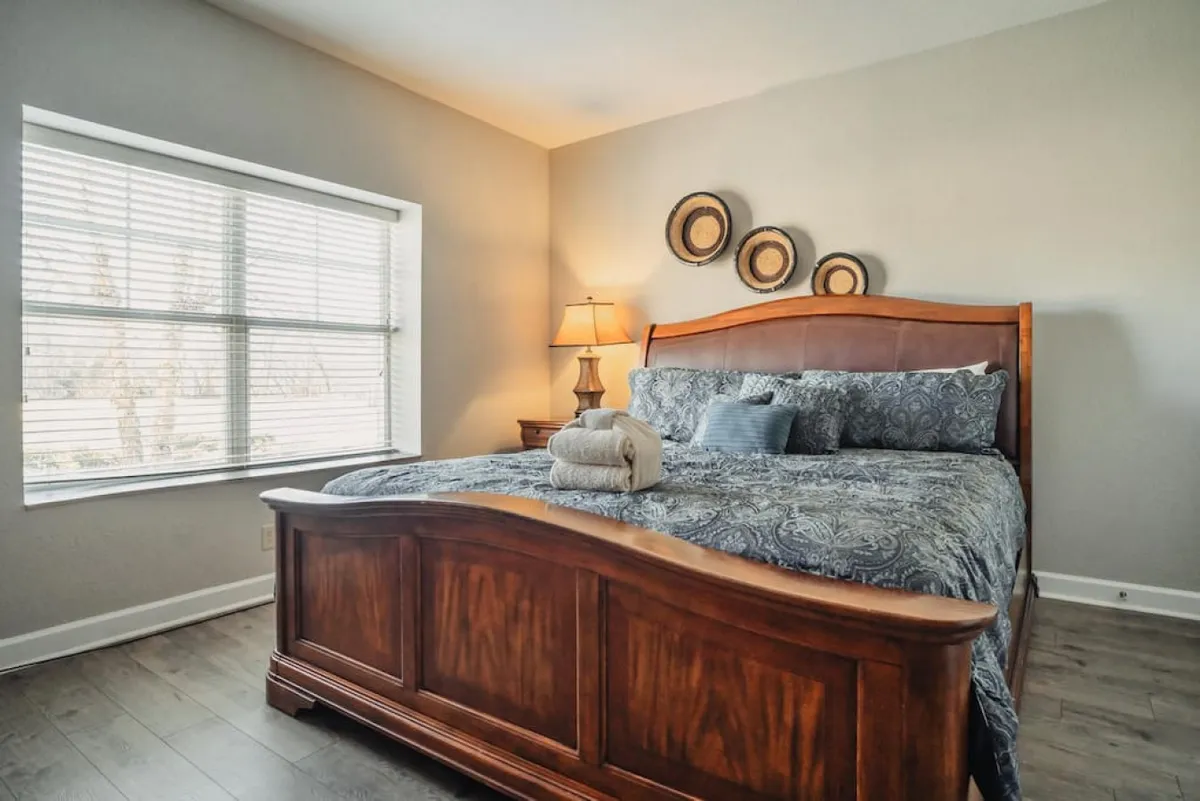 The image showcases a serene bedroom interior featuring a king-sized bed adorned with plush decorative pillows, a softly glowing table lamp, and a window gently streaming in natural light through white blinds. The scene exudes warmth and comfort, inviting relaxation and peaceful moments such as reading or unwinding after a day of adventure.