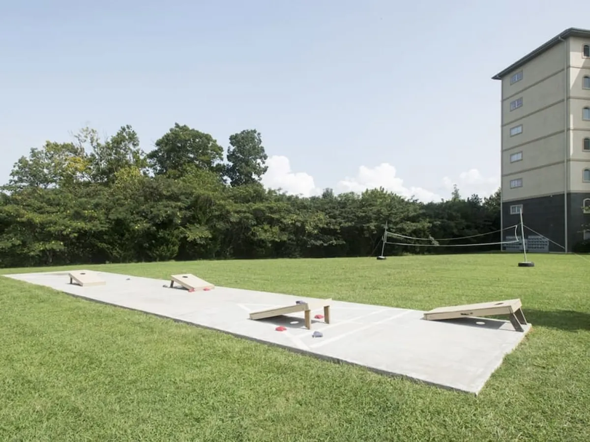 The image showcases a grassy outdoor recreation area with cornhole boards, a volleyball net, and a building in the background. It exudes a sense of community fun, outdoor games, and active engagement, inviting both kids and adults to play.