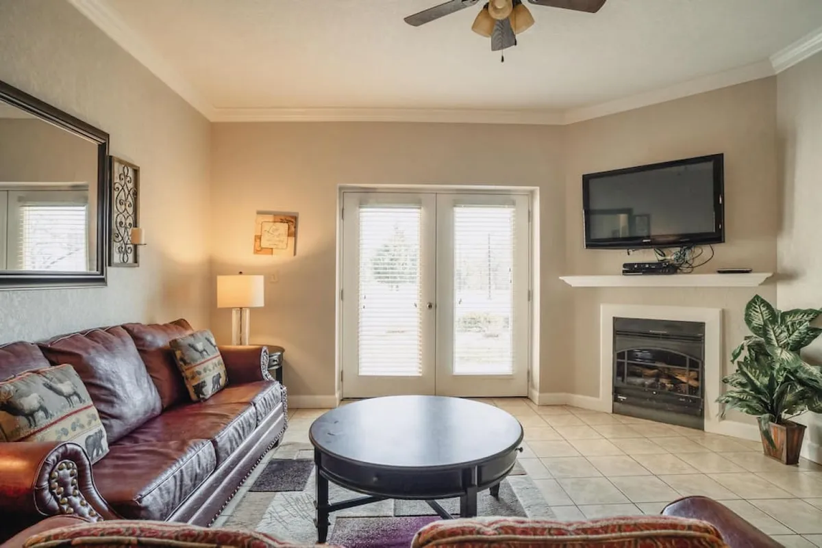 A warm, open-plan living area featuring a plush leather sofa, coffee table, large flat-screen TV, and a cozy fireplace. French doors let in natural light and offer a glimpse of the outdoor scenery, enhancing the welcoming ambiance.