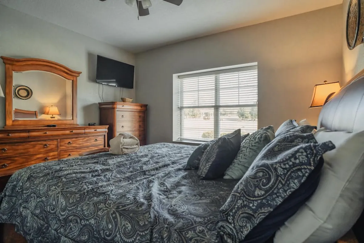 A cozy bedroom featuring a comfortable bed with decorative pillows, a wooden dresser, a wall-mounted TV, and a window with blinds. The mood is restful and modern, indicating amenities like in-room entertainment and a soothing retreat.