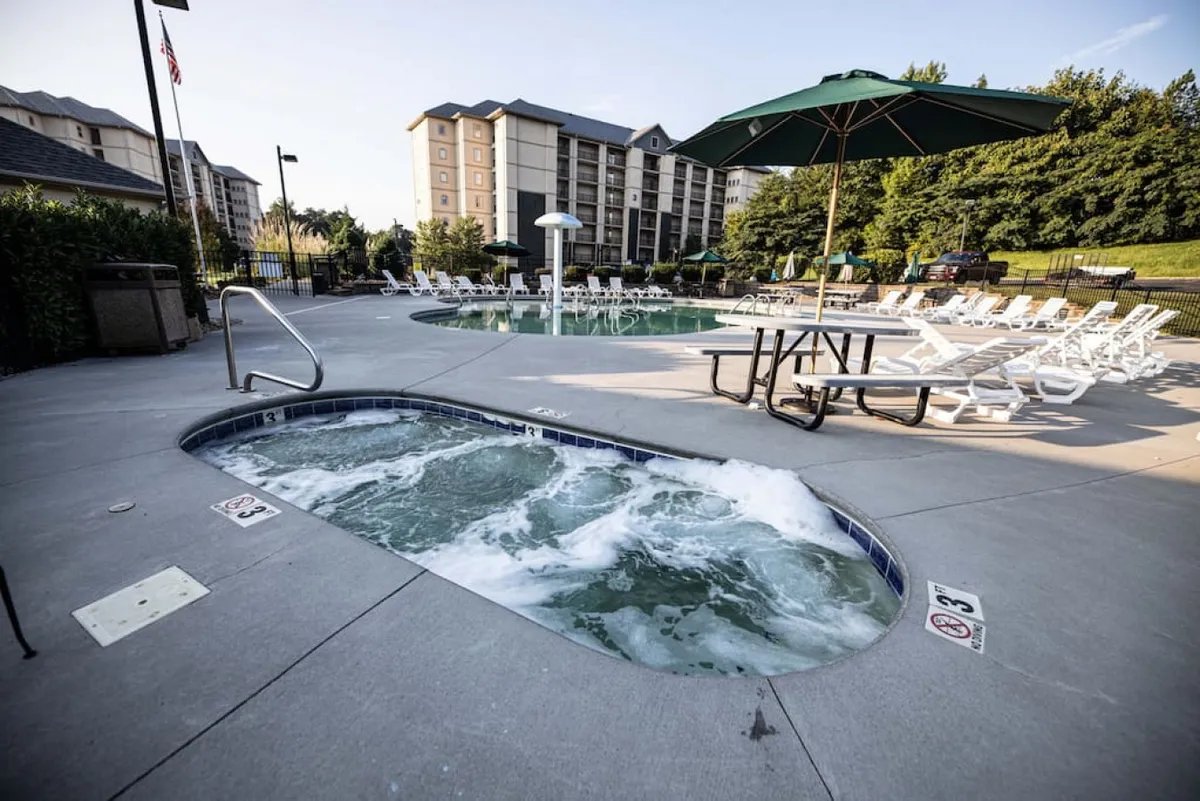An inviting resort pool area featuring a hot tub, swimming pool, lounge chairs, and a parasol. The space is designed for relaxation and socializing, suitable for guests of varying ages and abilities.