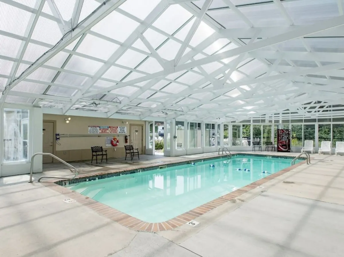 This image features a bright indoor swimming pool area with a glass ceiling that lets in plenty of natural light. Comfortable lounge chairs and poolside seating are arranged along the edges, creating a welcoming and lively environment for swimming, relaxing, and family gatherings.