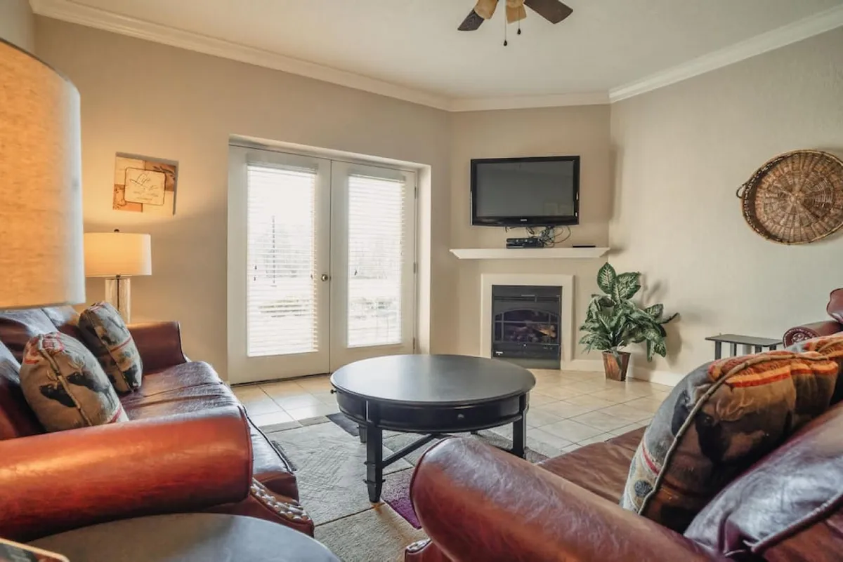 A cozy living room with leather sofas, a round coffee table, a fireplace, television, and sliding doors. The layout feels spacious and welcoming, ideal for gathering with friends or unwinding with a movie after exploring.